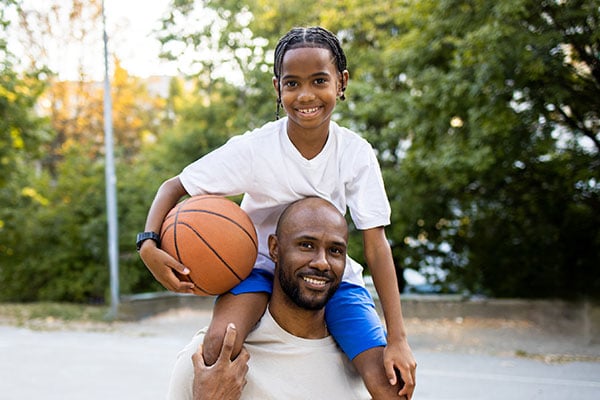 Latin American man carrying his son on shoulders while playing basketball at the outdoor basketball court.